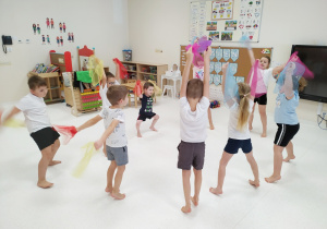 Dzieci stoją w kole, ćwiczą, trzymając w rękach kolorowe szyfonowe chustki.